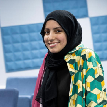 A student wearing a headscarf smiles at the camera with a blue and white chequered background.