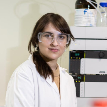 A student wearing safety goggles and a white lab coat smiles to the camera.