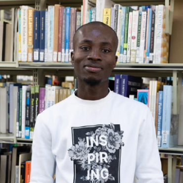 A student with a white top smiles at the camera with bookshelves behind them.