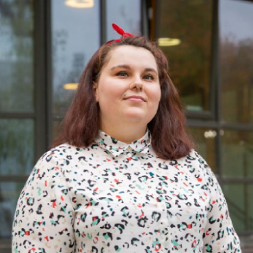 A student with a patterned shirt smiles to the camera with large windows behind them.