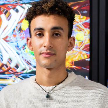 A student with dark hair and a white jumper looks into the camera with a diagram of a human body behind him.