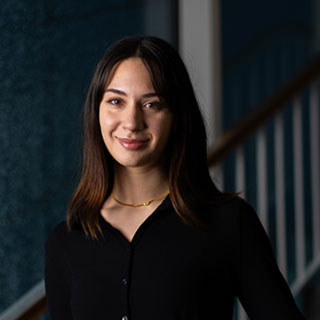 A head and shoulders image of a smiling person looking at the camera, standing at the bottom of a staircase.