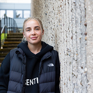 Student wearing casual clothing leaning against a building and smiles at camera