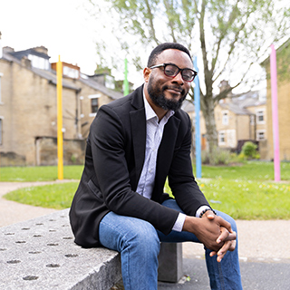 smartly dressed student wearing jeans, shirt and blazer smiles at the camera whilst sitting on a concrete bench with multicoloured poles in the background