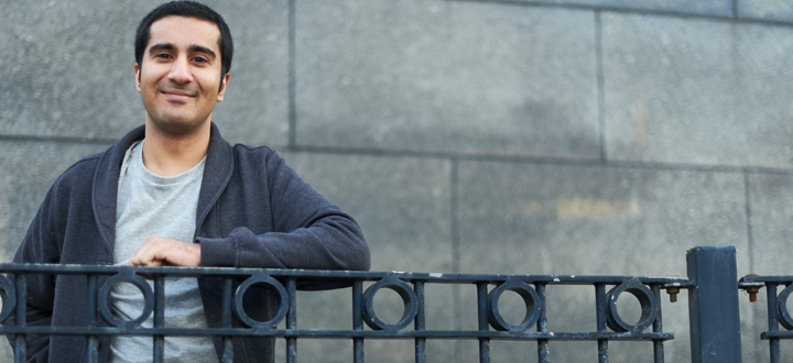 A person wearing a grey t-shirt with a dark cardigan standing in front of a brick wall and leaning on a rail